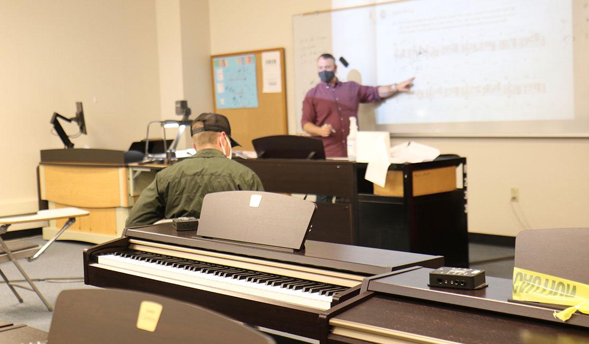 A masked instructor teaches students in a music classroom.