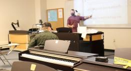 A masked instructor teaches students in a music classroom.