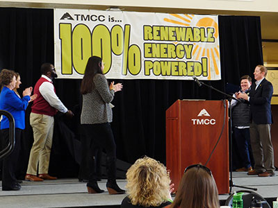 Staff members unroll a banner with the words TMCC is 100% renewable energy powered.
