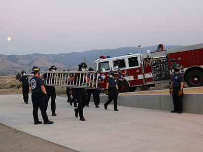 Cadets carrying a ladder with a fire truck in the background.
