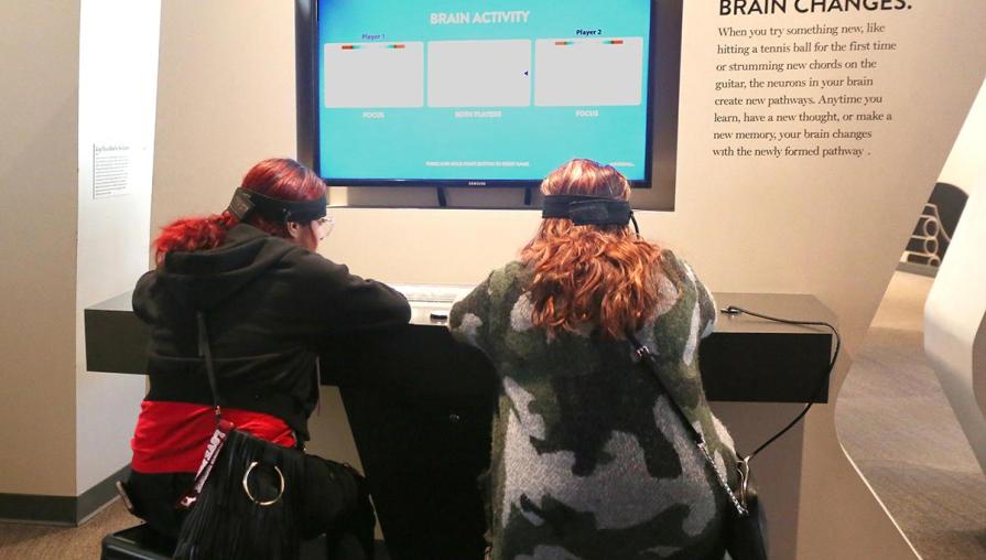 Two students wearing headbands are sat at a desk with a screen in front of them. 