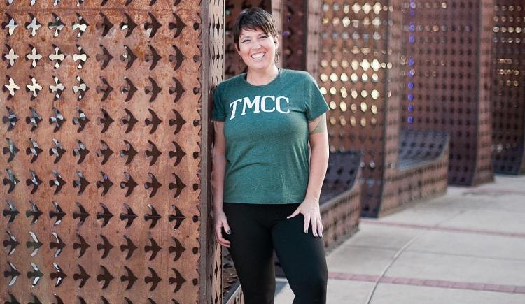 Monika Koshinski poses for a graduation photo outside, smiling and wearing a green TMCC t-shirt and black pants.