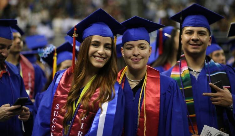 Three TMCC graduates donning blue caps and gowns with colorful sashes and cords gather excitedly for a picture in the Lawlor Events Center arena.