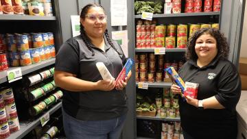 Two TMCC employees stand in a Wizard's Warehouse food pantry filled with canned meals, fruits, vegetables, and other donated items.