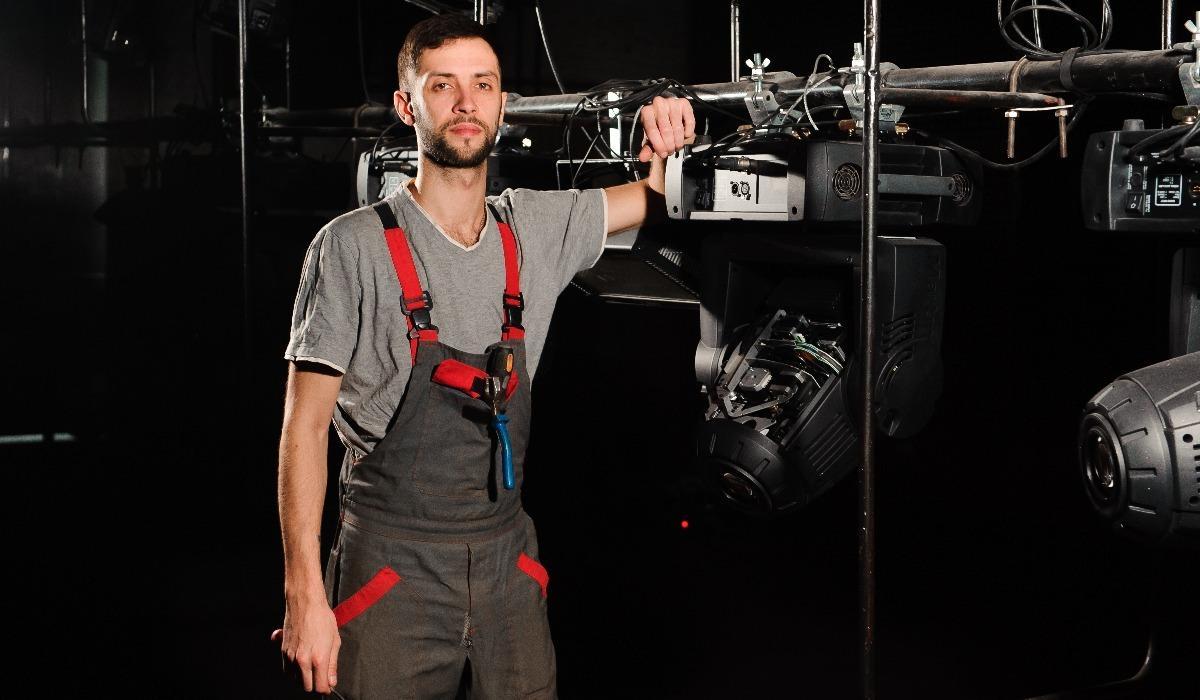 A theatre technician stands next to a lighting rig on a stage set.