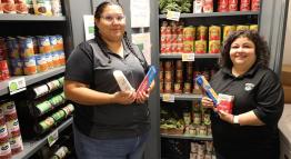 Two TMCC employees stand in a Wizard's Warehouse food pantry filled with canned meals, fruits, vegetables, and other donated items.